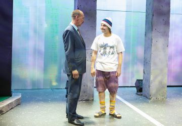 Kaksi miestä seisoo lavalla. Toisella on puku ja pukukengät, toisella T-paita, shortsit, värikkäät sukat, sandaalit ja pipo. Tausta on moderni, pastellivalaistus ja korkeat, kuvioidut paneelit.