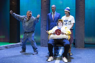 Neljä miestä on näyttämöllä teatteriesityksessä. Yksi farkkutakkinen mies elehtii tarmokkaasti, toinen seisoo puvussaan katselemassa, kun taas kolmas pipoon pukeutunut mies seisoo keltaiseen takkiin ja hattuun pukeutuneen istuvan miehen takana, jolla on punainen viuhka kädessään.