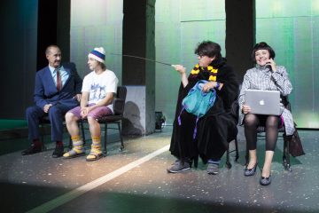 Lavalla istuu neljä ihmistä: mies puvussa, nuori mies t-paidassa ja shortseissa, henkilö mustassa kaapussa ja keltaisessa huivissa kepin kanssa ja nainen liikeasussa kannettavan tietokoneen ja puhelimen kanssa, kaikki teatteriympäristössä.