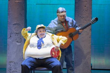 Kaksi näyttelijää esiintyy lavalla. Toinen on pukeutunut keltaiseen takkiin ja lippikseen, istuu purkki kädessään ja elehtii eloisasti. Toinen, farkkutakkiin ja otsapantaan pukeutunut, seisoo takana ja soittaa kitaraa. Sininen valaistus määrittää näyttämön.