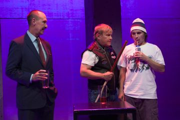 Kolme miestä seisoo yhdessä violetissa valaistuksessa; yhdellä pukumiehellä on kädessään juoma, toisella nahkaliiviin pukeutuneella on kädessään biljardikeppi, ja kolmas, jolla on pipo ja rento pukeutuminen, juo tölkistä.