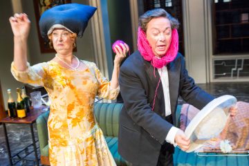 Keltaiseen mekkoon pukeutunut nainen, jolla on tyyny päässään, nostaa kätensä ylös, kun taas pukuun pukeutuneella miehellä on vaaleanpunaista lankaa kasvojensa ympärillä ja hänellä on hattu. Molemmat näyttävät olevan keskellä koomista kohtausta lavalla.