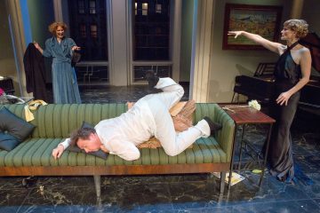 Lavalla on kolme ihmistä: valkoisiin pukeutunut mies makaa kasvot alaspäin vihreällä sohvalla, ja kaksi tyylikkääseen mekkoon pukeutunutta naista seisoo molemmin puolin ja elehtii hänelle eloisasti hyvin valaistussa, tyylikkäässä olohuoneessa.