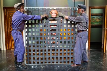 Kaksi muuttomiehiksi pukeutunutta miestä pitää pystyssä suurta kokoontaitettavaa sängynrunkoa, jonka sisällä on huoneen lavastusta muistuttavalla näyttämöllä kukkavaatteisiin pukeutunut nainen, joka on jäänyt loukkuun yllättyneenä.