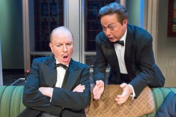 Kaksi smokkiin pukeutunutta miestä istuu vihreällä sohvalla; toisella on kädet ristissä ja hän näyttää järkyttyneeltä, kun taas toinen kumartuu eteenpäin ja elehtii vilkkaasti kuin yrittäisi selittää tai suostutella häntä.