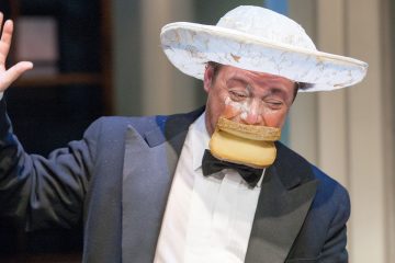 Smokkiin ja suureen valkoiseen hattuun pukeutuneella henkilöllä on valkoista kermaa kasvoillaan ja leipäviipale suussaan, ja hän nostaa yhden kätensä leikkisästi tai koomisesti.