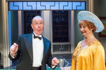 Smokkiin pukeutunut vanhempi mies tekee hölmöjä ilmeitä seisoessaan keltaiseen mekkoon ja suureen siniseen hattuun pukeutuneen hymyilevän vanhemman naisen vieressä kirkkaasti valaistussa lavasteessa.