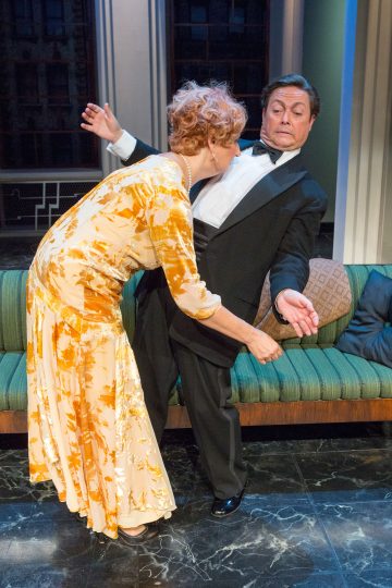 Keltaiseen kukkamekkoon pukeutunut nainen nojaa kohti mustaan smokkiin pukeutunutta, hämmästyneenä taaksepäin kumartuvaa miestä vihreällä vintage-sohvalla sijaitsevassa teatterikohtauksessa.