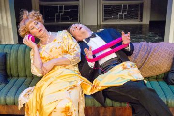 Kaksi juhlapukuista ihmistä istuu vihreällä sohvalla. Keltaiseen mekkoon pukeutunut nainen pitää vaaleanpunaista lankaa korvallaan, silmät kiinni. Smokkipukuinen mies nojaa taaksepäin silmät kiinni, lanka käsien välissä.