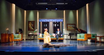 Kirkkaasti valaistu näyttämö, jossa on tyylikäs olohuone, jossa on art deco -huonekaluja, maalauksia ja flyygeli. Kaksi näyttelijää, toinen istumassa ja toinen seisomassa, ovat näyttämön keskellä, ja etualalla on tumma turkismatto.