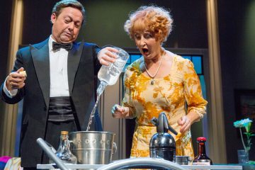 Smokkiin pukeutunut mies kaataa suuren pullon kirkasta nestettä metalliseen ämpäriin, kun keltaiseen kukkamekkoon pukeutunut yllättynyt nainen katselee. He seisovat pöydän ääressä, jossa on pulloja ja juomien valmistusvälineitä.