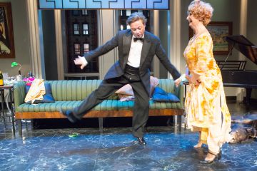 Smokkiin pukeutunut mies tanssii energisesti hymyilevän, keltaiseen mekkoon pukeutuneen naisen kanssa olohuoneessa, jonka taustalla on vihreä sohva ja piano.