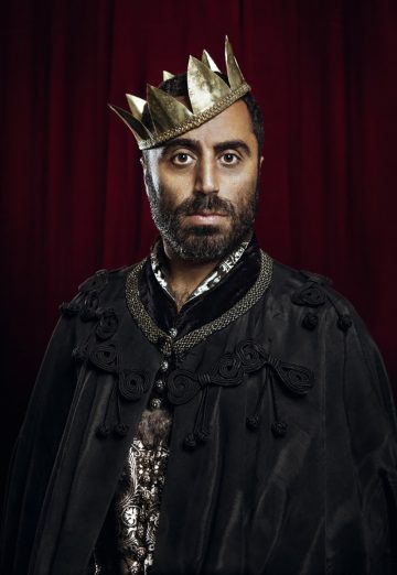 Parrakas mies, jolla on kultainen kruunu ja koristeellinen musta viitta, seisoo syvän punaisen verhon edessä ja katsoo suoraan kameraan.