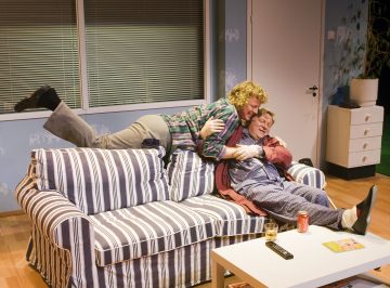 Kaksi ihmistä istuu raidallisella sohvalla olohuoneessa; toinen pyjama-asuinen henkilö istuu pystyssä, kun taas toinen, vapaa-ajan vaatteissa, makaa leikkisästi heidän päällään, molemmat hymyillen. Heidän edessään on pöytä, jossa on juomia ja kaukosäädin.