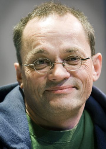 Keski-ikäisellä henkilöllä, jolla on lyhyet ruskeat hiukset, pyöreät silmälasit ja hento hymy, on vihreä paita ja tumma takki. Tausta on pehmeästi sumea.