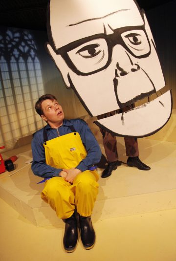 Keltaisiin haalareihin ja mustiin saappaisiin pukeutunut mies istuu lattialla ja katsoo ylöspäin suurta, kuvitettua ja silmälasipäistä henkilön kasvojen leikkausta, jota joku hänen takanaan seisova henkilö pitää. Kohtaus vaikuttaa teatraaliselta ja ilmeikkäältä.