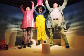 Kolme värikkäisiin sadetakkeihin pukeutunutta ihmistä nostaa iloisesti käsiään, kun vesi roiskuu alas heidän yläpuolellaan olevasta vaaleanpunaisesta ämpäristä, ja he seisovat kirkkaasti valaistulla näyttämöllä, jonka ympärillä on leikkisää rekvisiittaa.