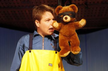 Siniseen takkiin ja keltaiseen esiliinaan pukeutunut henkilö pitelee ruskeaa nallekarhua, jolla on musta silmäside, ja katsoo sitä suu auki.