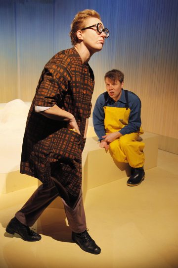 Kaksi ihmistä esiintyy lavalla. Toinen, jolla on ruskeat kuviolliset vaatteet ja suuret pyöreät silmälasit, kävelee eteenpäin, kun taas toinen, jolla on sininen paita ja keltainen haalari, istuu pölkyn päällä ja katsoo heitä. Tausta on minimalistinen ja pehmeästi valaistu.