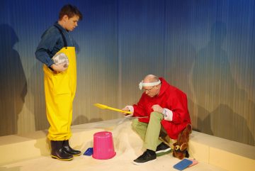 Lavalla on kaksi ihmistä; toisella on pallo kädessään ja keltainen haalari, toinen istuu lelulapio ja -ämpäri kädessään, punainen paita ja otsapanta yllään, nallen ja lelulapion vieressä kirkkaasti valaistussa ympäristössä.