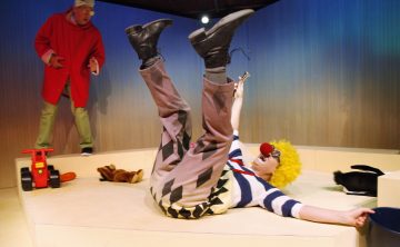 Klovni, jolla on keltainen peruukki, punainen nenä ja raidallinen paita, makaa selällään jalat koholla ja pitää kädessään pientä esinettä. Toinen punatakkinen esiintyjä elehtii vilkkaasti taustalla. Leluja on hajallaan lähistöllä.