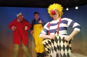 Lavalla kolme esiintyjää: yksi punatakissa ja päähineessä, toinen keltaisessa haalarissa osoittamassa, ja klovni raidallisessa paidassa, henkselihousuissa ja keltaisessa kiharassa peruukissa, jossa on punainen nenä, pitelee työkalua ja poseeraa hassusti.