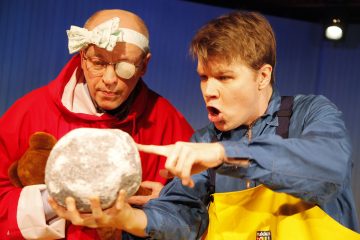 Kaksi värikkäisiin asuihin pukeutunutta miestä katselee harmaata, pyöreää esinettä; toinen osoittaa sitä yllättyneenä, kun taas toinen, jolla on silmälasit, päässään sideharso ja silmälappu, pitää nallea kädessään ja katselee tarkasti.