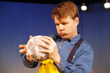 Siniseen paitaan ja keltaiseen esiliinaan pukeutunut mies tutkii kirkkaassa näyttämövalaistuksessa intensiivisesti pyöreää, harmaata, kuvioitua esinettä molemmin käsin.