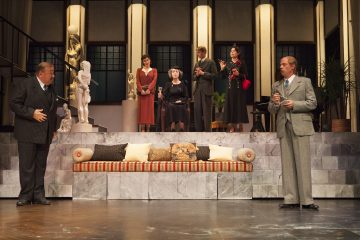 Seitsemän näyttelijää vanhoissa juhlavaatteissa esiintyy lavasteissa, jotka muistuttavat tyylikästä olohuonetta, jossa on marmoriportaat, veistoksia ja raidallinen sohva. Ryhmä näyttää käyvän dramaattista keskustelua.