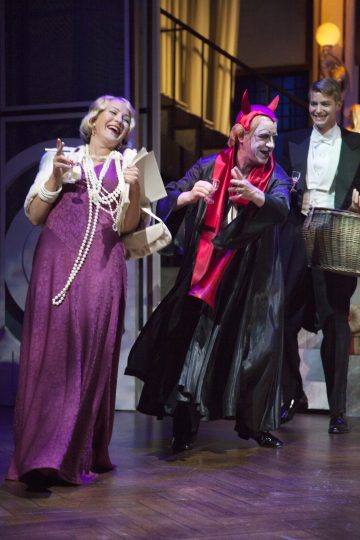 Violettiin mekkoon ja pitkiin helmiin pukeutunut nainen nauraa, kun paholaispukuun ja meikkiin pukeutunut henkilö elehtii dramaattisesti hänen vieressään lavalla, ja taustalla hymyilee virka-asuun pukeutunut mies, joka pitää koria kädessään.