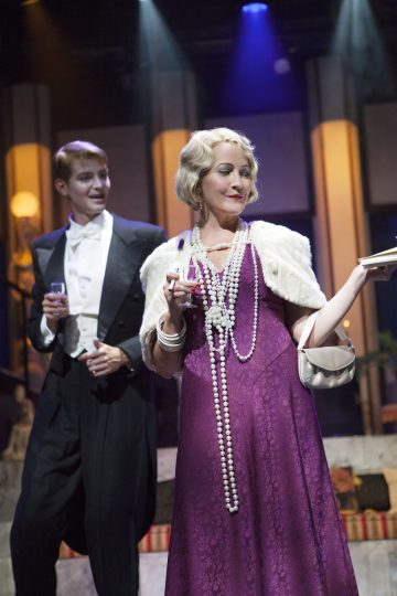 Kaksi 1920-luvun tyyliin pukeutunutta näyttelijää seisoo näyttämön valojen alla. Pitkään violettiin mekkoon, helmikaulakoruihin ja valkoiseen huiviin pukeutunut nainen pitelee juomaa ja lautasta. Hänen takanaan olevalla miehellä on smokki, ja hänellä on myös juoma kädessään.