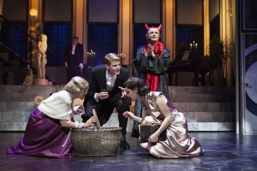 Neljä virallisesti pukeutunutta näyttelijää kerääntyy näyttämöllä korin ympärille, joista yhdellä on paholaisen sarvet ja punainen viitta. Kohtaus sijoittuu tyylikkääseen, kynttilöiden valossa valaistuun huoneeseen, jossa on marmoriportaat ja klassinen sisustus.