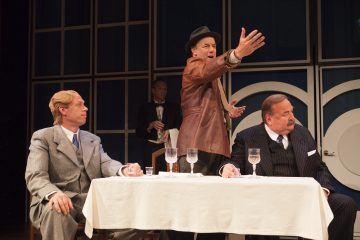 Kolme pukumiestä istuu pöydän ääressä, jossa on valkoinen pöytäliina ja lasit, ja heidän takanaan seisoo hattupäinen ja ruskeatakkinen mies, joka elehtii dramaattisesti. Toinen mies seisoo taustalla lautasliina kädessään.