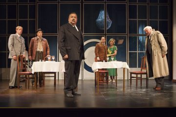 Kuusi vintage-asuihin pukeutunutta näyttelijää esiintyy lavalla, jonka yläpuolella on kaksi ruokapöytää, tuolit ja kattokruunu. Taustalla on suuret ikkunat. Keskeinen näyttelijä seisoo eteenpäin ja hallitsee huomiota.