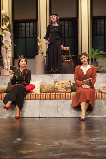 Kolme vintage-tyylisiin mekkoihin pukeutunutta naista on lavastettu tyylikkääseen olohuoneeseen, jossa on patsaita, kasveja ja kuviollisia tyynyjä. Kaksi istuu matalalla marmoriseinällä, kun taas yksi seisoo heidän takanaan dramaattisen näköisenä ja keppi kädessään.