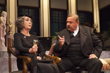 Mustiin vaatteisiin ja hattuun pukeutunut vanhempi nainen pitelee pientä esinettä istuessaan pukumiehen vieressä, joka elehtii kädellään; molemmat näyttävät käyvän vakavaa keskustelua lavalla.