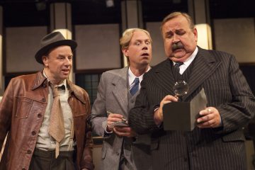 Kolme vanhaan pukuun pukeutunutta miestä seisoo yhdessä; yksi pitää suurennuslasia mustan laatikon yllä, toinen tekee muistiinpanoja, ja kolmas katsoo hämmästyneenä. Lavasteena näyttää olevan näyttämö, jossa on dramaattinen valaistus.