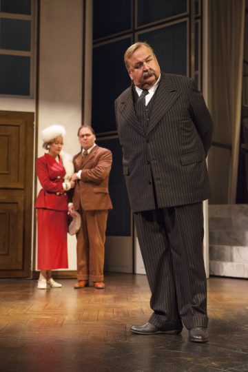 Etualalla seisoo nauhamaiseen pukuun pukeutunut mies kädet selän takana ja näyttää vakavalta. Taustalla punapukuinen nainen ja ruskeapukuinen mies tarkkailevat häntä lavasteissa.