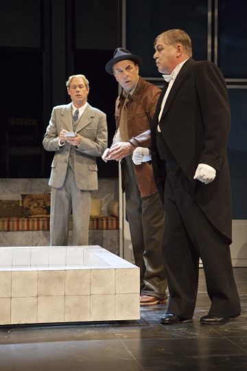 Kolme miestä seisoo sisätiloissa keskustellen. Yhdellä on vaalea puku, toisella ruskea nahkatakki ja hattu ja kolmannella musta frakkipuku ja valkoiset käsineet. He vaikuttavat keskittyneiltä ja tarkkaavaisilta. Taustalla on sohva.