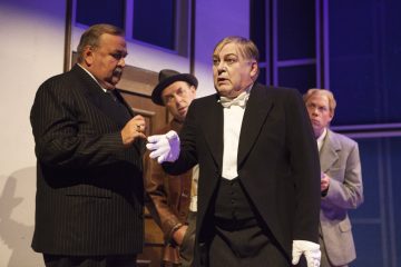 Neljä vanhanaikaisiin pukuihin ja asuihin pukeutunutta miestä seisoo dramaattisessa valaistuksessa; yhdellä on valkoiset hansikkaat ja hän näyttää puhuvan, kun taas muut kuuntelevat tarkkaavaisesti ja vakavin ilmein.