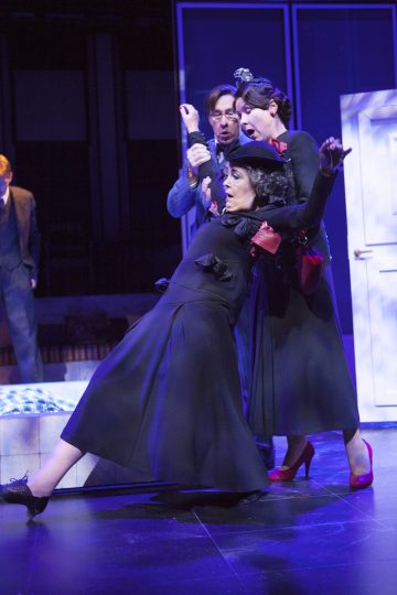 Kolme näyttelijää näyttämöllä dramaattisessa keskitoiminnassa: yksi mustapukuinen nainen, jolla on leveälippainen hattu, nojaa taaksepäin, toinen nainen, jolla on musta mekko ja punaiset korkokengät, ja mies seisoo heidän takanaan pitelemässä naista kädestä. Sänky ja ovi ovat lähellä.