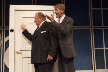 Kaksi pukumiestä seisoo näyttämöllä valkoisen oven vieressä. Toinen vanhemmista miehistä katsoo ovea kohti käsi koholla, kun taas hänen takanaan oleva nuorempi mies elehtii innostuneesti, mikä viittaa siihen, että he näyttelevät dramaattisessa tai komediallisessa teatterikohtauksessa.