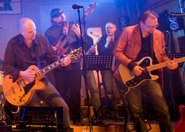 Lavalla esiintyy viiden miehen yhtye, jossa kaksi kitaristia on edessä, basisti ja kosketinsoittaja takana ja rumpali osittain näkyvissä. Taustalla värikkäät lavavalot ja musiikkivälineet.