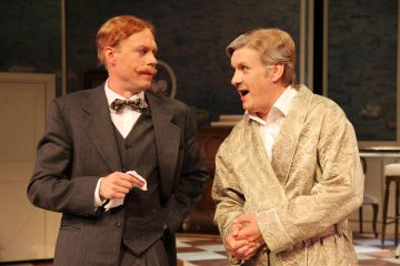 Kaksi vanhempaa, aikakauden pukuihin pukeutunutta miestä keskustelee sisätiloissa; toisella on tumma puku ja rusetti, ja hänellä on nenäliina kädessään, kun taas toisella on kuviollinen kaapu ja hän elehtii eloisasti. Ympäristö vaikuttaa näyttämönäytelmältä.