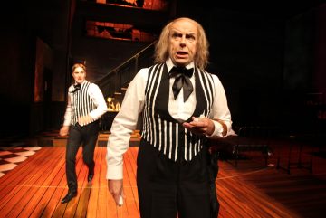 Kaksi mustavalkoraidallisiin raidallisiin liiveihin ja rusetteihin pukeutunutta näyttelijää esiintyy hämärästi valaistulla lavalla, jossa on puulattia; toinen seisoo etualalla ja näyttää yllättyneeltä, kun taas toinen liikkuu taustalla lähellä portaikkoa.