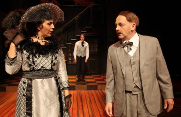 Kaksi 1900-luvun alun pukuihin pukeutunutta näyttelijää kohtaa toisensa näyttämöllä; nainen, jolla on höyhenpukuinen hattu ja ruudullinen mekko, ja mies, jolla on harmaa puku. Kolmas henkilö seisoo taustalla portaikon vieressä.