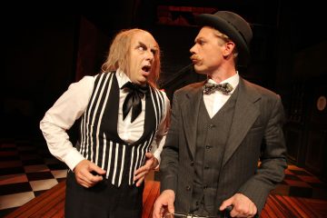 Lavalla on kaksi vintage-asuihin pukeutunutta näyttelijää. Toinen, jolla on villi harmaa tukka ja raidallinen liivi, näyttää järkyttyneeltä. Toinen, puku ja keilahattu, näyttää vakavalta. He seisovat ruudullisella lattialla dramaattisessa valaistuksessa.