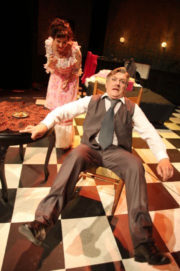 Pukumies kaatuu yllättyneen näköisenä tuolille mustavalkoruudulliselle lattialle. Vaaleanpunaiseen kukkamekkoon pukeutunut nainen seisoo hänen takanaan ja vaikuttaa huolestuneelta. Kohtaus sijoittuu hämärästi valaistuun huoneeseen.