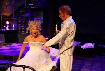 Valkoiseen mekkoon pukeutunut nainen katsoo järkyttyneenä, kun raidalliseen pyjamaan pukeutunut mies vetää lakanaa taaksepäin hämärässä huoneessa, jossa on dramaattinen näyttämövalaistus ja tumma, teatraalinen lavastus.