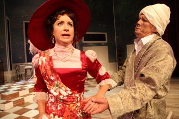 Punaisessa aikakauden puvussa ja suuressa hatussa oleva nainen näyttää ilmeikkäältä, kun taas koristeltuihin kaapuihin ja valkoiseen päähineeseen pukeutunut mies pitelee hellästi naisen käsivartta näyttämöllä, jossa on ruudullinen lattia.